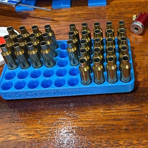 Pode incluir: Um suporte de cartuchos de plástico azul preenchido com estojos de balas de latão. O suporte está rotulado como "6.5 Creedmoor". Componentes adicionais de plástico azul são visíveis no fundo, juntamente com uma ferramenta vermelha e prateada.