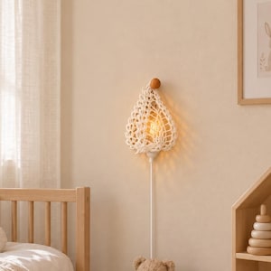 Op de afbeelding: Een wandlamp met een crèmekleurige macramé kap met een open vlechtpatroon, opgehangen aan een kleine houten knop. De lamp verspreidt een warm licht van een zichtbare gloeilamp en hangt aan een wit snoer tegen een effen muur.