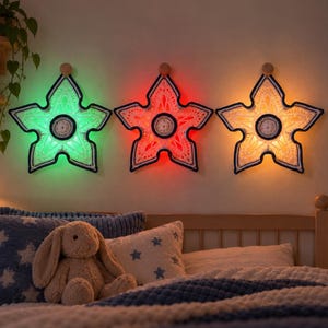 May include: Three star-shaped wall hangings featuring intricate white crochet patterns with dark blue borders. Each star is illuminated from behind, emitting green, red, and warm yellow light respectively. They are mounted on a wall above a bed.