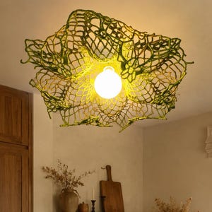 May include: A ceiling light fixture featuring a green, open-work crocheted shade with an irregular, organic shape. The shade surrounds a single, glowing white spherical light bulb that casts a warm, bright light downward into the room.