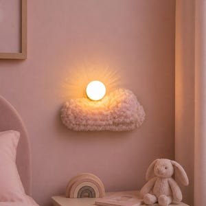 May include: A wall-mounted light fitting shaped like a fluffy, textured cloud in a soft pink hue. The light features a central, glowing circular bulb that emits a warm, diffused light, creating a sunburst effect against the wall surface.
