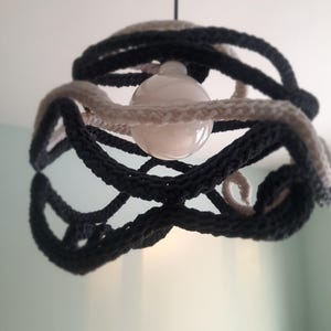 May include: A modern pendant light fixture with a spherical bulb at the center. The shade is made of intertwined, thick, knitted rope in shades of gray and off-white. The light hangs from a black cord.