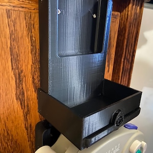 Puede incluir: Soporte de pared negro impreso en 3D para un mando de Nintendo GameCube. El soporte tiene una bandeja rectangular y una ranura para sujetar el mando. El mando es gris con botones de colores y el logotipo de Nintendo GameCube.