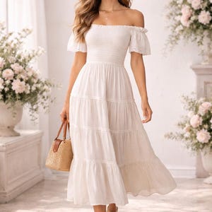 Może przedstawiać: Biała sukienka midi z odkrytymi ramionami, falbanowymi rękawami i warstwową spódnicą. Sukienka jest połączona z beżowymi sandałami i tkana torebką z brązową skórzaną rączką. Tło przedstawia kompozycje kwiatowe i okno.