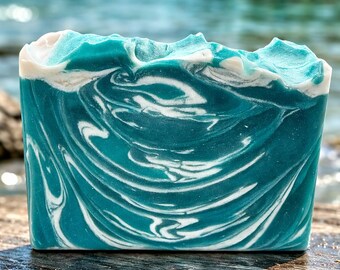 May include: A rectangular bar of handmade soap with a teal and white swirled design, resembling ocean waves. The top edge has a textured, wave-like appearance. The soap is set against a blurred background of water and a distant shoreline.