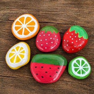 May include: Six hand-painted rocks in the shape of fruit slices. The collection includes an orange, lemon, lime, watermelon, and two strawberries. Each rock is painted with bright colors and white accents, set against a wooden background.