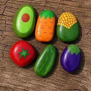 Może przedstawiać: Ręcznie malowane kamienie dekoracyjne w kształcie warzyw. Kolekcja obejmuje awokado, marchewkę, kukurydzę, pomidora, ogórka i bakłażana. Każdy kamień jest pomalowany żywymi kolorami i szczegółami.