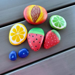 May include: A collection of hand-painted rocks depicting various fruits. The assortment includes a peach, lime, lemon, watermelon, strawberry, and two blueberries. Each rock is painted with vibrant colors and detailed designs, creating a playful and artistic display.