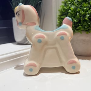 May include: A ceramic rocking horse figurine in a pastel color palette. The horse is primarily white with pink and blue accents on its mane, tail, and body. The figurine has a whimsical, vintage aesthetic.