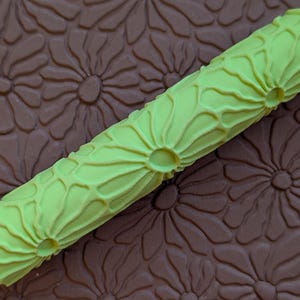 May include: A close-up of a light green rolling pin with a daisy pattern. The rolling pin is cylindrical and has a textured surface. The background is a brown surface with a repeating daisy pattern.