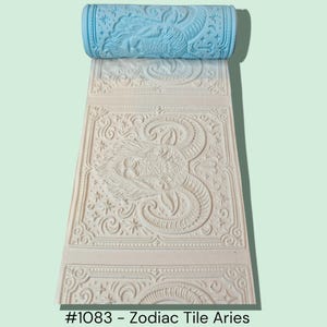 May include: A light blue roller with an Aries zodiac design, rolled over a cream-colored surface with the same embossed Aries pattern. The design includes the text "#1083 - Zodiac Tile Aries".