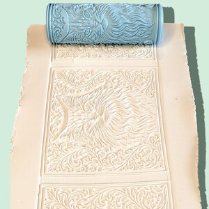 May include: A light blue cylindrical patterned roller resting on a flat white surface. The roller features an embossed lion face surrounded by intricate floral scrollwork, which is imprinted into the white material beneath the roller.