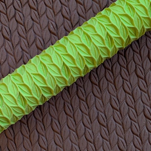 May include: A lime green rolling pin with a leaf pattern is positioned diagonally across a brown surface with a matching leaf pattern. The rolling pin's design is raised, creating a textured effect. The background is a darker shade of brown.