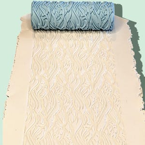 May include: A light blue cylindrical patterned texture roller resting on a flat, off-white rectangular slab of material. The roller features a repeating embossed design of flowing, organic lines that are imprinted into the surface below.