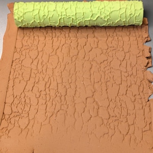 May include: A lime green textured rolling pin sits on a sheet of brown clay. The clay has been rolled to create a cracked, textured surface. The rolling pin is cylindrical and has a repeating pattern.