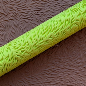May include: A close-up of a textured, lime green roller with a pattern resembling grass. The roller is positioned diagonally against a brown, textured surface, creating a contrast in color and texture. The image highlights the detailed design.