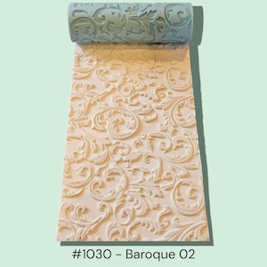 May include: A light blue patterned rolling pin positioned above a white rectangular slab of clay. The clay features a raised, embossed scrollwork design that matches the pattern on the roller. The text #1030 - Baroque 02 is printed below.
