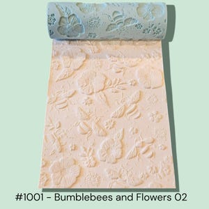 May include: A light blue rolling pin with a floral and bee pattern is unrolled onto a cream-colored surface. The pattern includes flowers, leaves, and bumblebees. The text at the bottom reads "#1001 - Bumblebees and Flowers O2."