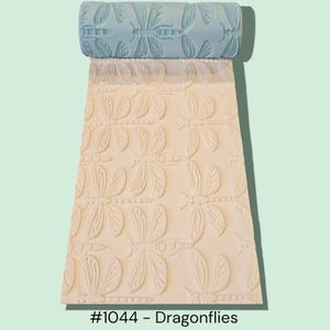 May include: A light blue cylindrical rolling pin with a debossed dragonfly pattern, shown pressing into a flat, off-white clay surface that displays the same repeating dragonfly design. The text #1044 - Dragonflies is printed at the bottom.
