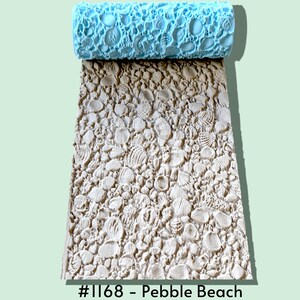 May include: A light blue cylindrical texture roller with a raised pattern of various seashells and pebbles. The roller is positioned above a rectangular beige clay impression showing the same detailed seashell and pebble design with the text #1168 - Pebble Beach.