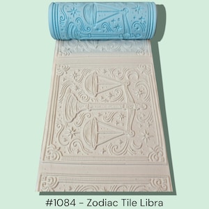 May include: A light blue roller and a beige tile with a Libra zodiac design. The roller has a repeating pattern of the Libra symbol, stars, and swirls. The tile shows the embossed design of the Libra symbol, stars, and decorative elements. The text reads "#1084 - Zodiac Tile Libra."