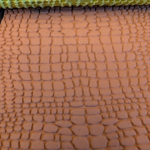 May include: A textured, brown clay surface with a repeating pattern of rectangular shapes, resembling crocodile skin. A lime green roller with a honeycomb pattern is positioned above the clay, suggesting a tool for imprinting the design.