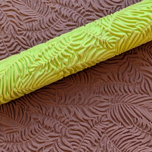 May include: A close-up of a lime green textured roller with a leaf pattern, resting on a brown surface with a matching leaf pattern. The roller is cylindrical and appears to be made of rubber or a similar material.
