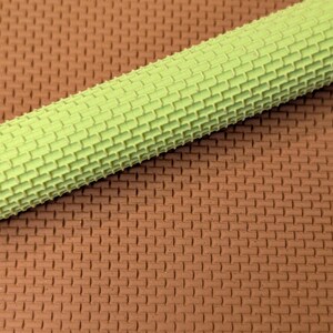 May include: A close-up shot of a light green rolling pin with a brick pattern. The rolling pin is positioned diagonally across a brown surface, also with a brick pattern.