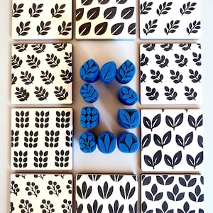 May include: A collection of eight square white tiles featuring various black leaf and berry patterns, arranged around a central cluster of nine blue foam stamps, each carved with distinct botanical shapes and leaf motifs.