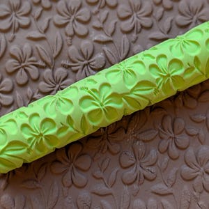May include: A lime green rolling pin with a floral pattern is positioned diagonally across a brown surface with a matching floral design. The rolling pin's design features detailed flower and leaf impressions, creating a textured effect. The brown surface also has a floral pattern.