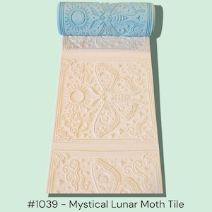 May include: A light blue roller with a sun and moth design, rolled over a cream-colored surface with a repeating pattern. The surface features a large moth and sun design. The text "#1039 - Mystical Lunar Moth Tile" is at the bottom.