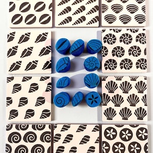 May include: A set of twelve square ceramic coasters featuring black seashell patterns on a white background, arranged in a grid around a central cluster of ten small, blue, foam seashell-shaped stamps used for crafting.