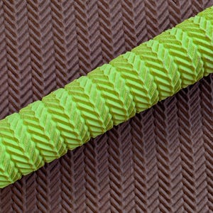 May include: Close-up of a textured, neon green grip with a repeating chevron pattern. The grip is cylindrical and lies diagonally across a brown surface with a matching chevron design. The grip is likely for a sports equipment handle.