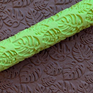May include: A lime green rolling pin with a leaf pattern rests on a brown surface with a matching leaf pattern. The rolling pin is cylindrical and features a repeating design of tropical leaves. The brown surface has the same leaf pattern.