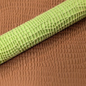 May include: A close-up of a textured, light green rolling pin with a grid pattern, resting on a brown, textured surface. The rolling pin is cylindrical and appears to be made of a flexible material.