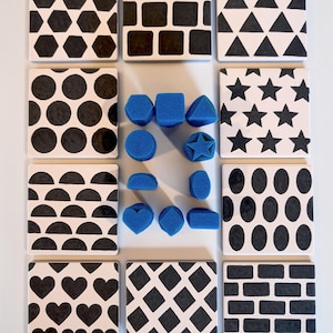 May include: A set of twelve square white tiles featuring various black geometric patterns including hexagons, squares, triangles, circles, stars, semicircles, ovals, hearts, diamonds, and bricks, accompanied by a collection of small blue foam shapes.