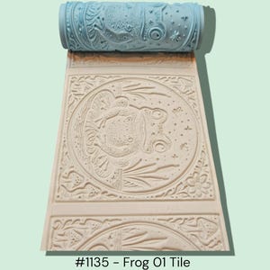 May include: A light blue textured roller with a frog design, used for creating embossed patterns. The roller is shown with a sample of the embossed pattern in off-white, featuring a frog and floral elements. Text at the bottom reads "#1135 - Frog 01 Tile."