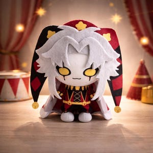 Puede incluir: Muñeco de peluche con disfraz de bufón, con cara blanca, ojos amarillos y pelo blanco. El muñeco viste un atuendo rojo y negro con detalles dorados y un sombrero a juego con cascabeles dorados. Decoración de circo en el fondo.