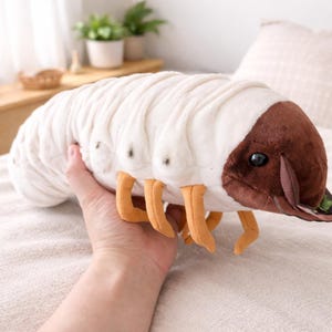 Può includere: Un peluche a forma di larva, con corpo bianco, testa marrone e zampe arancioni. Il giocattolo morbido è tenuto in mano, mostrando le sue dimensioni e il suo design. La larva ha dettagli a punti neri. Giocattolo per bambini.