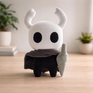Pode incluir: Um brinquedo de pelúcia de um personagem com cabeça branca, olhos pretos grandes e chifres curvos. Ele usa um cachecol cinza e uma capa preta, segurando uma espada cinza. O brinquedo está em uma superfície de madeira.