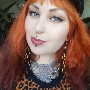 May include: A woman with bright red hair wearing a black hat, a leopard print top, and long gold earrings. She has multiple facial piercings, including a septum ring and a lip ring.