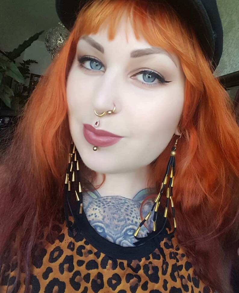 May include: A woman with bright red hair wearing a black hat, a leopard print top, and long gold earrings. She has multiple facial piercings, including a septum ring and a lip ring.