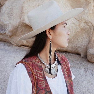 May include: A woman wearing a white cowboy hat, a white t-shirt, a red and black patterned vest, and long silver earrings. She is also wearing a silver necklace with a feather pendant.