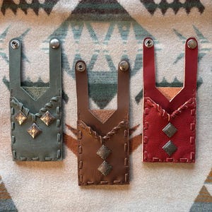 May include: Three leather phone cases in teal, brown, and red, each with a unique design. The cases feature decorative stitching, metal studs, and a strap with a snap closure. They are displayed on a patterned textile.