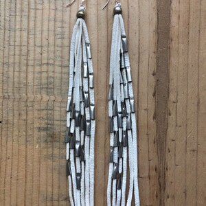 May include: White leather earrings with silver metal accents. The earrings are long and dangle, with multiple strands of leather and metal beads.