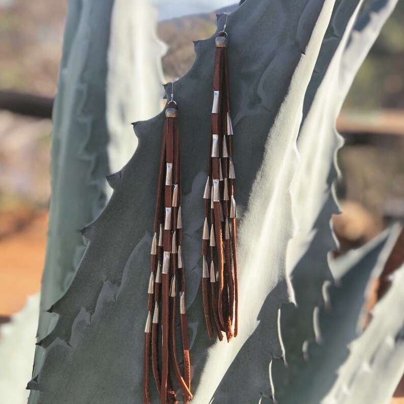 May include: A pair of long brown leather earrings with silver accents. The earrings are hanging from a plant.