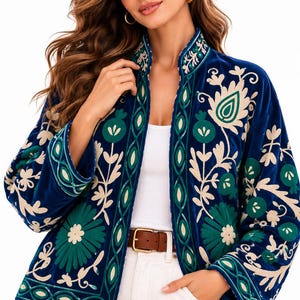 May include: A deep blue velvet jacket featuring intricate embroidery in shades of cream and forest green. The design includes floral and botanical motifs along the front panels, collar, and sleeves, with a structured open front and long sleeves.