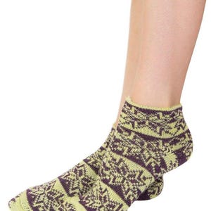 Puede incluir: Calcetines tobilleros con un patrón geométrico. Los calcetines son principalmente de color verde claro con un patrón morado oscuro. Se ven en los pies de una persona, con las piernas visibles. Hechos de material de punto.