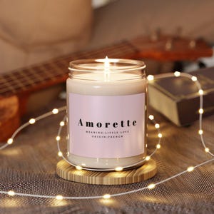 May include: A lit candle in a glass jar with a pink label that reads "Amorette, meaning little love, origin: French." The candle sits on a small wooden coaster, surrounded by string lights. A guitar and book are in the background.