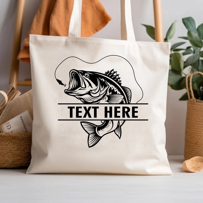 May include: A cream-colored tote bag featuring a black and white illustration of a largemouth bass with an open mouth, a fishing hook, and the text "TEXT HERE" in a bold font. The bag has two cream-colored handles.